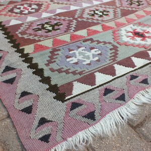 May include: A woven rug with a geometric pattern in shades of pink, gray, brown, and white. The rug features repeating diamond and floral motifs, with a fringed edge. The design is symmetrical and the rug appears to be made of wool.