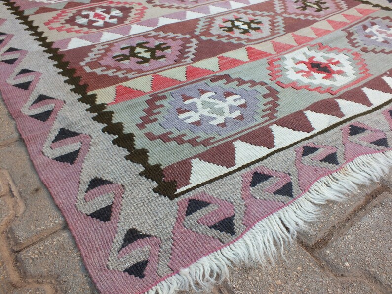 May include: A woven rug with a geometric pattern in shades of pink, gray, brown, and white. The rug features repeating diamond and floral motifs, with a fringed edge. The design is symmetrical and the rug appears to be made of wool.