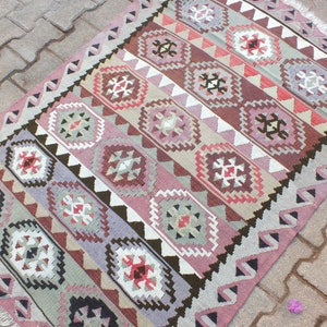 May include: A rectangular, patterned rug with a geometric design. The rug features a repeating pattern of diamond-shaped medallions in shades of pink, brown, and gray, with white and black accents. The rug has a fringed edge and measures approximately 4 x 6 feet.