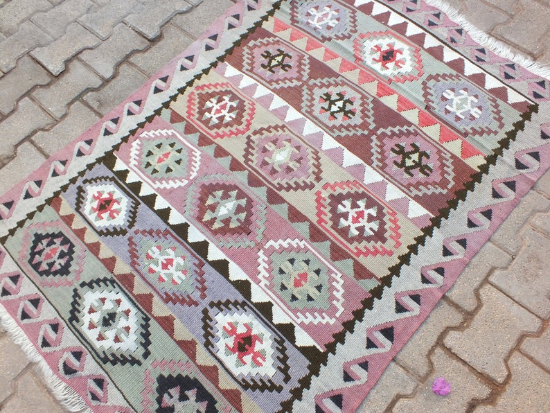 May include: A rectangular, patterned rug with a geometric design. The rug features a repeating pattern of diamond-shaped medallions in shades of pink, brown, and gray, with white and black accents. The rug has a fringed edge and measures approximately 4 x 6 feet.