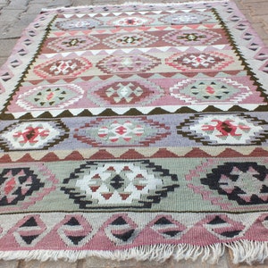 May include: A rectangular, hand-woven rug with a geometric pattern. The rug features a repeating design of diamond and oval shapes in shades of pink, purple, brown, and green. The edges are finished with a fringe.