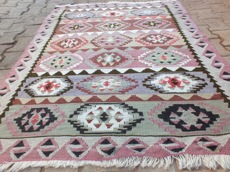 May include: A rectangular, hand-woven rug with a geometric pattern. The rug features a repeating design of diamond and oval shapes in shades of pink, purple, brown, and green. The edges are finished with a fringe.