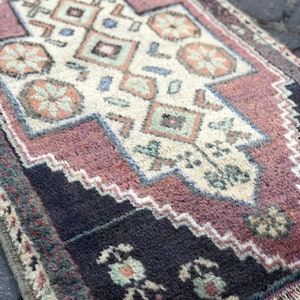 Op de afbeelding: Een klein, rechthoekig tapijt met een ingewikkeld geometrisch patroon. Het tapijt heeft een cr&egrave;mekleurig middenveld met herhalende vormen in tinten oranje, blauw en groen. De rand is diepzwart met bloemmotieven en een gefranjerde rand.