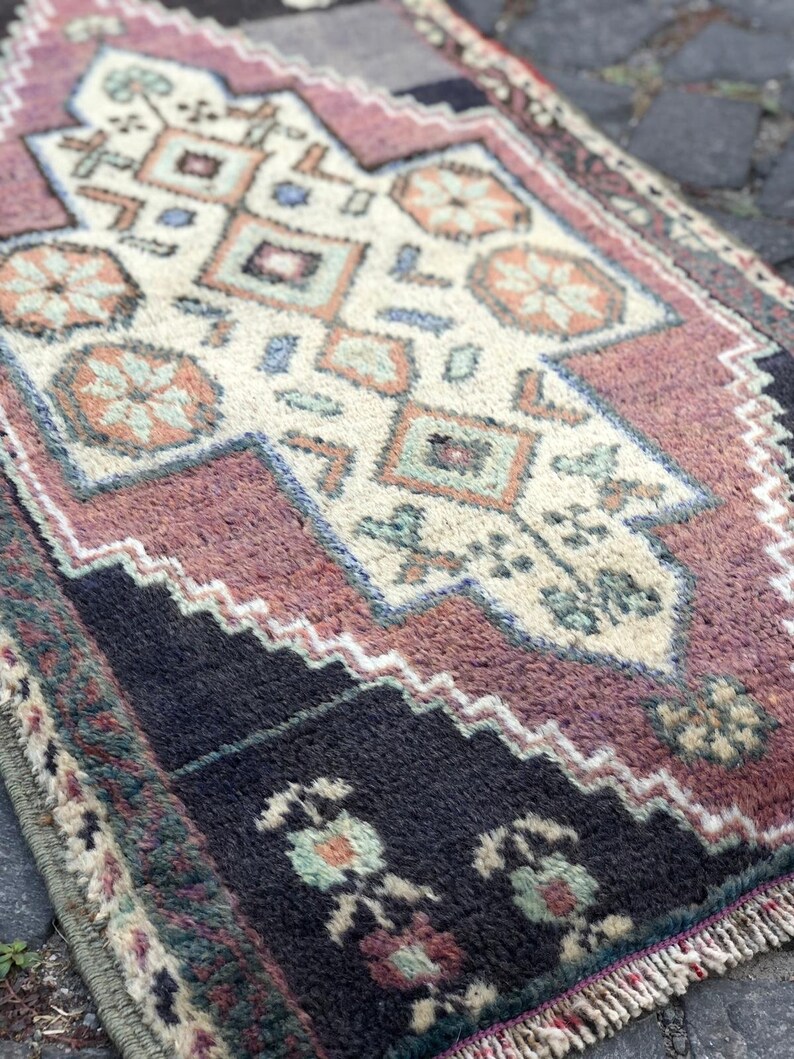 Op de afbeelding: Een klein, rechthoekig tapijt met een ingewikkeld geometrisch patroon. Het tapijt heeft een cr&egrave;mekleurig middenveld met herhalende vormen in tinten oranje, blauw en groen. De rand is diepzwart met bloemmotieven en een gefranjerde rand.