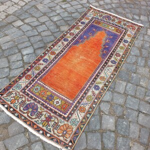May include: A vintage orange prayer rug with a traditional floral design and a blue and white prayer niche. The rug has a white fringe border.