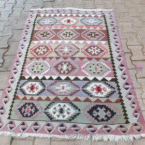 May include: A rectangular, hand-woven rug with a geometric pattern. The rug features a repeating design of diamond and oval shapes in shades of pink, brown, gray, and white. The edges are trimmed with a fringe.