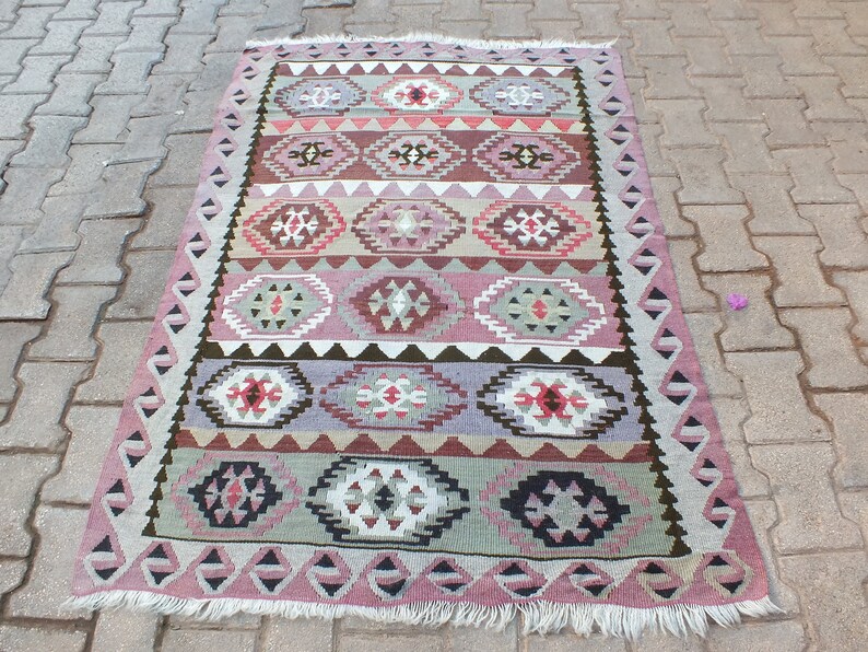 May include: A rectangular, hand-woven rug with a geometric pattern. The rug features a repeating design of diamond and oval shapes in shades of pink, brown, gray, and white. The edges are trimmed with a fringe.