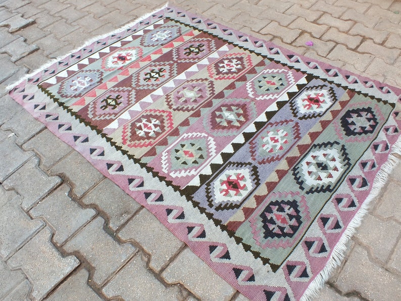May include: A rectangular, hand-woven rug with a geometric pattern. The rug features a repeating design of diamond and rectangular shapes in shades of pink, brown, gray, and white. The edges are finished with a fringe.