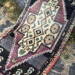 Op de afbeelding: Een klein, rechthoekig tapijt met een centraal geometrisch ontwerp in cr&egrave;me, bordeauxrood en blauwgroen. Het tapijt heeft een zwarte rand met bloemaccenten en een gefranjerde rand. Het tapijt ligt op een stenen oppervlak.