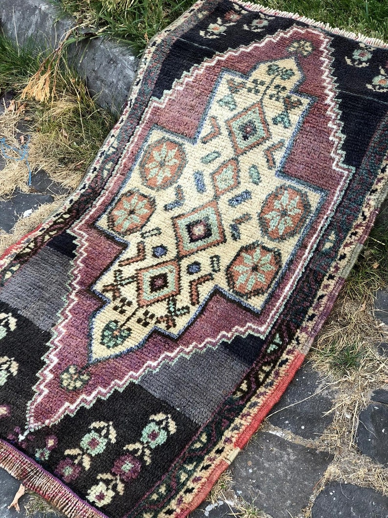 Op de afbeelding: Een klein, rechthoekig tapijt met een centraal geometrisch ontwerp in cr&egrave;me, bordeauxrood en blauwgroen. Het tapijt heeft een zwarte rand met bloemaccenten en een gefranjerde rand. Het tapijt ligt op een stenen oppervlak.