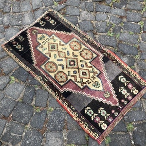 Op de afbeelding: Een rechthoekig, vintage tapijt met een centraal geometrisch ontwerp in cr&egrave;me, bordeauxrood en zwart. Het tapijt heeft bloemen- en geometrische patronen, met een gefranjerde rand. Het tapijt ligt op een kasseien oppervlak.