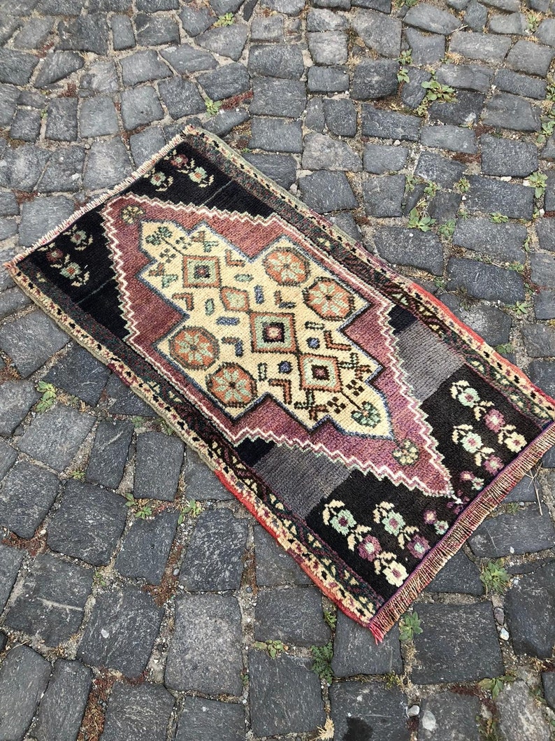 Op de afbeelding: Een rechthoekig, vintage tapijt met een centraal geometrisch ontwerp in cr&egrave;me, bordeauxrood en zwart. Het tapijt heeft bloemen- en geometrische patronen, met een gefranjerde rand. Het tapijt ligt op een kasseien oppervlak.