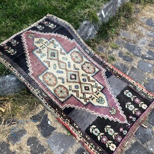 Op de afbeelding: Een rechthoekig, handgeweven tapijt met een centraal geometrisch ontwerp in cr&egrave;me, paars en blauw. Het tapijt heeft zwarte randen met bloempatronen en gefranjerde randen. Het tapijt ligt op een stenen oppervlak.