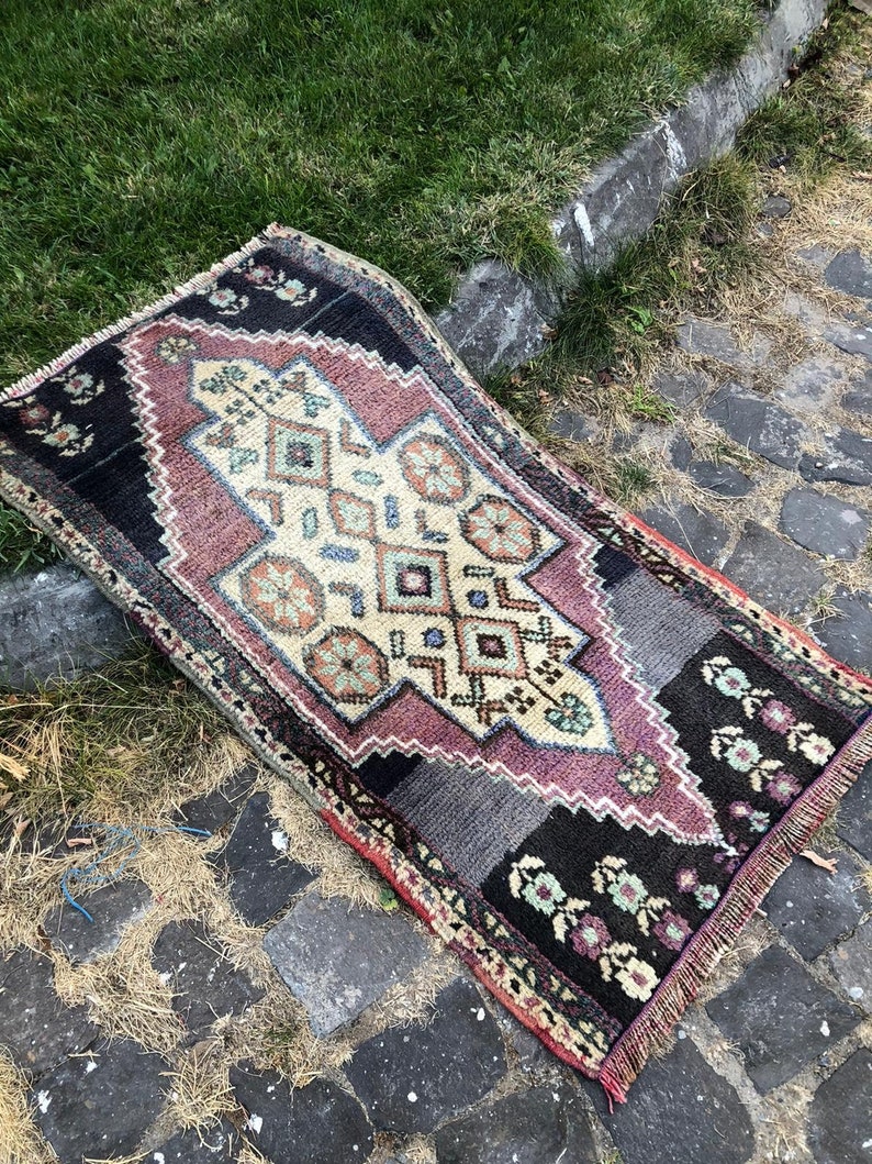 Op de afbeelding: Een rechthoekig, handgeweven tapijt met een centraal geometrisch ontwerp in cr&egrave;me, paars en blauw. Het tapijt heeft zwarte randen met bloempatronen en gefranjerde randen. Het tapijt ligt op een stenen oppervlak.