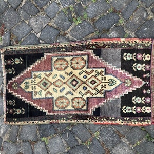 Op de afbeelding: Een klein, rechthoekig tapijt met een geometrisch patroon. Het tapijt heeft een centrale ruitvorm in beige, bordeauxrood en zwart, met bloemaccenten en een donkere rand. Het ligt op een kasseienoppervlak.