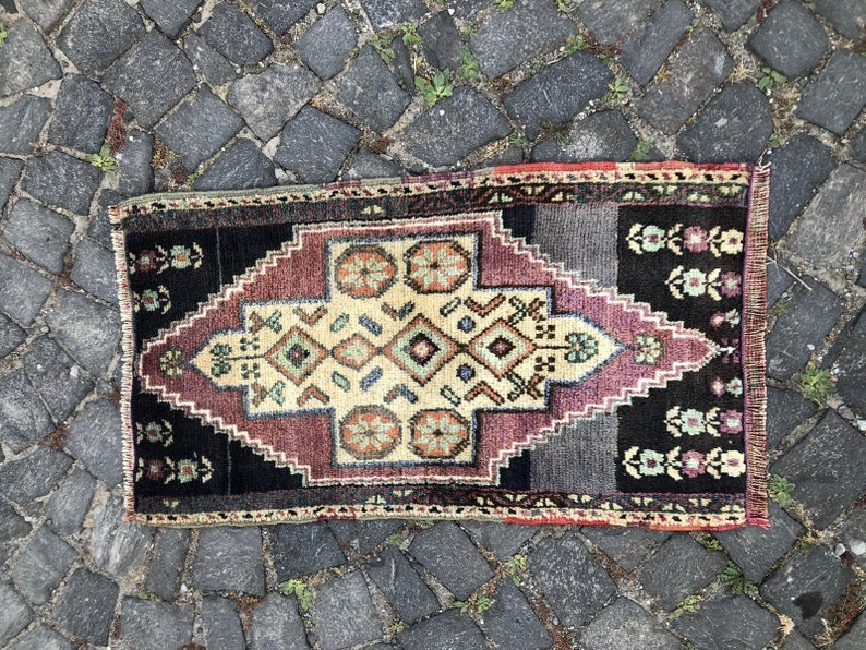 Op de afbeelding: Een klein, rechthoekig tapijt met een geometrisch patroon. Het tapijt heeft een centrale ruitvorm in beige, bordeauxrood en zwart, met bloemaccenten en een donkere rand. Het ligt op een kasseienoppervlak.