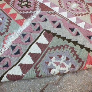 May include: A colorful, patterned rug with a folded edge. The rug features geometric designs in shades of pink, gray, red, and white. The rug's fringe is visible along the edge. The rug is likely made of woven fabric.