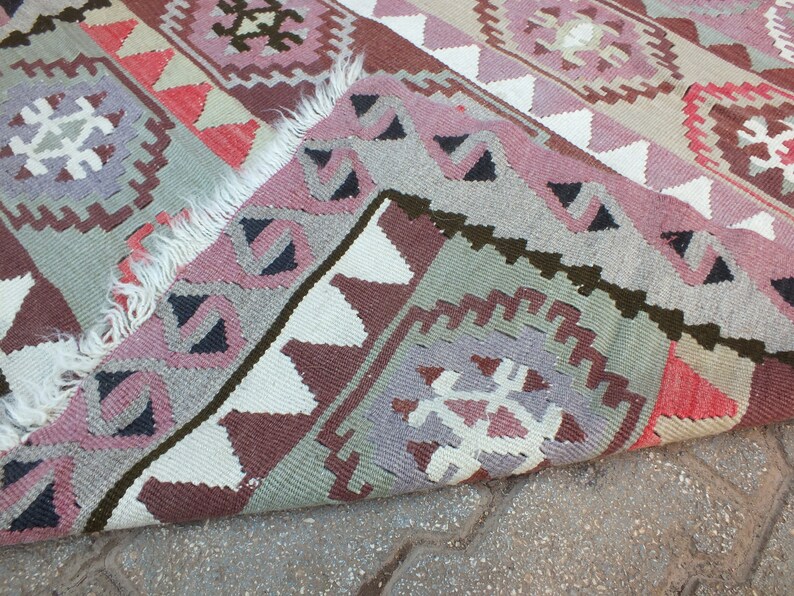 May include: A colorful, patterned rug with a folded edge. The rug features geometric designs in shades of pink, gray, red, and white. The rug's fringe is visible along the edge. The rug is likely made of woven fabric.