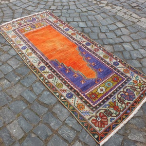 May include: A traditional Turkish prayer rug with a vibrant orange center and a purple border. The rug has intricate floral and geometric patterns in shades of red, blue, yellow, and green. The rug is fringed on all sides.