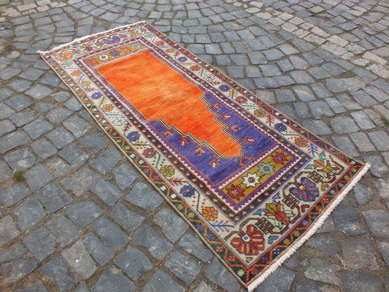 May include: A traditional Turkish prayer rug with a vibrant orange center and a purple border. The rug has intricate floral and geometric patterns in shades of red, blue, yellow, and green. The rug is fringed on all sides.