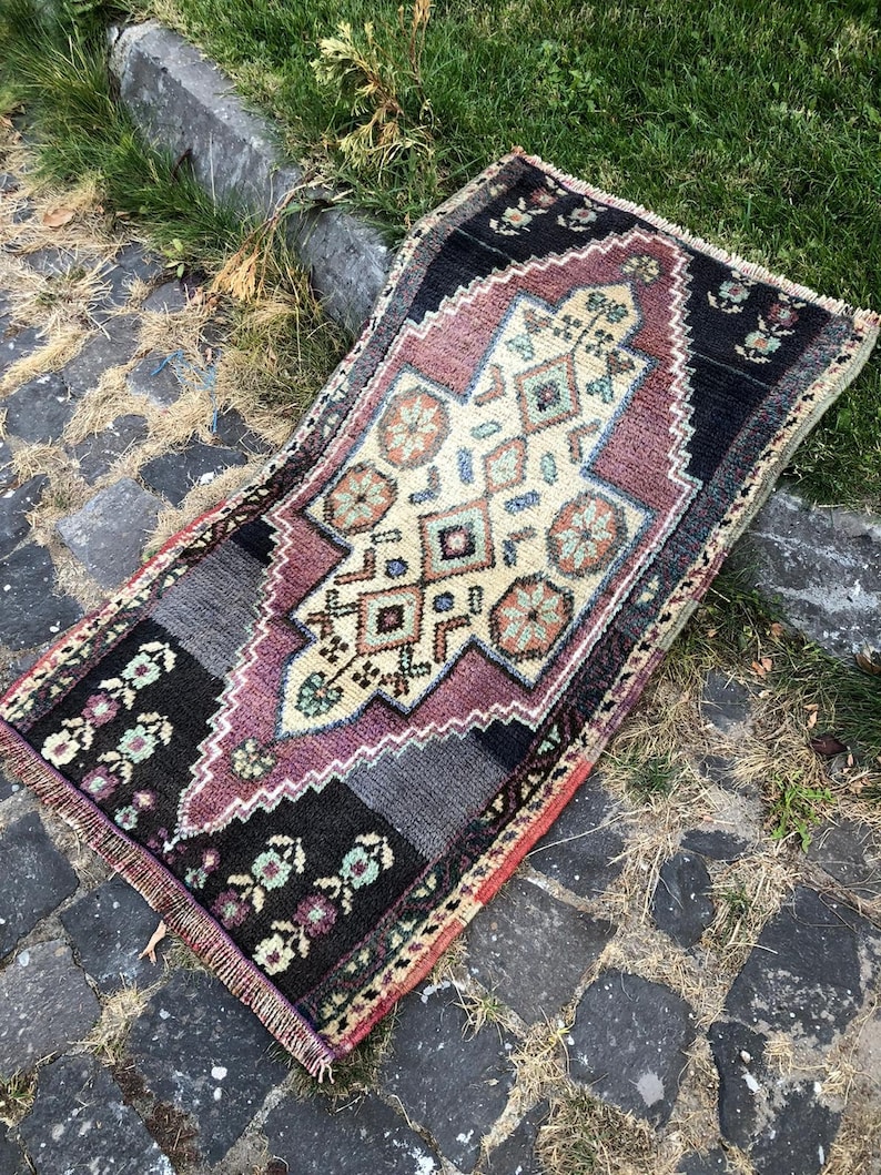 Op de afbeelding: Een rechthoekig, handgeweven tapijt met een centraal geometrisch ontwerp in cr&egrave;me, paars en teal. Het tapijt heeft een zwarte rand met bloempatronen en wordt buiten op een geplaveide ondergrond getoond.