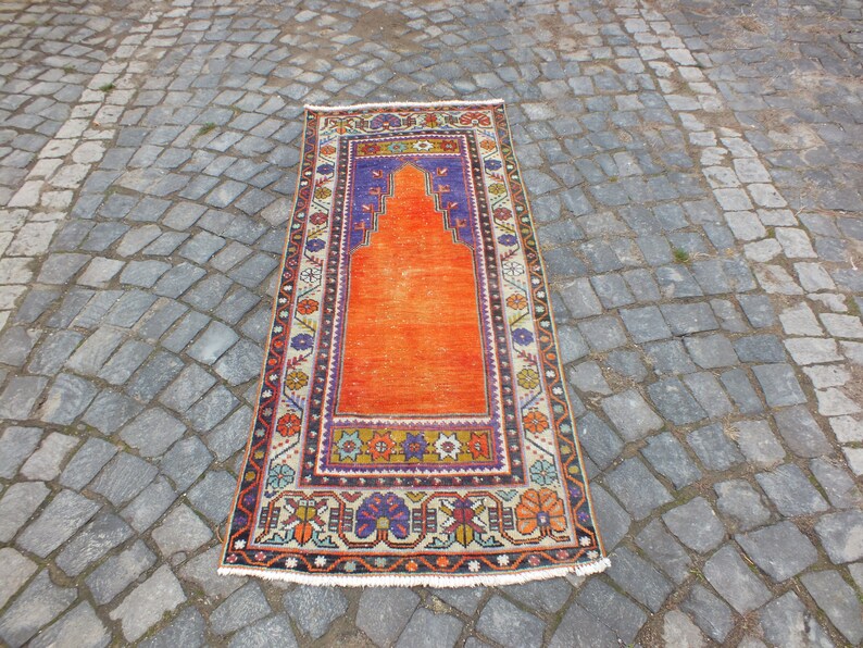 May include: A traditional orange prayer rug with a colorful floral border and a central arch design. The rug has a fringed edge and is made of wool.