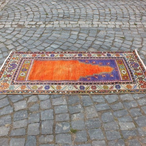 May include: A traditional orange and purple prayer rug with intricate floral patterns and a central design. The rug is laid out on a gray cobblestone surface.