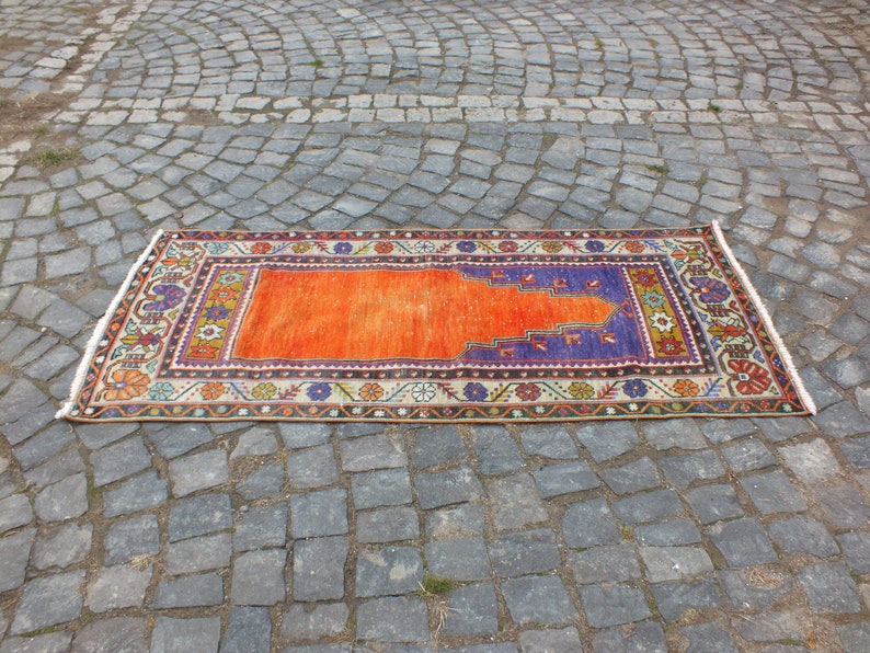 May include: A traditional orange and purple prayer rug with intricate floral patterns and a central design. The rug is laid out on a gray cobblestone surface.