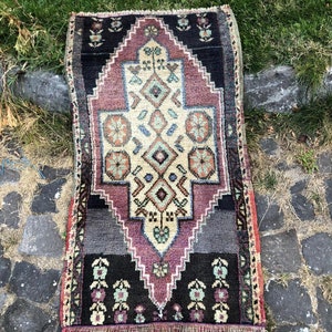 Op de afbeelding: Een rechthoekig, handgeweven tapijt met een centraal geometrisch ontwerp in cr&egrave;me, paars en zwart. Het tapijt heeft een gedetailleerd patroon met bloemen- en ruitmotieven en een gefranjerde rand. Het tapijt is ongeveer 61 cm lang en 30 cm breed.