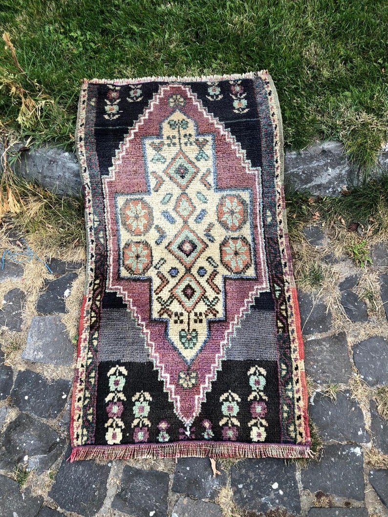 Op de afbeelding: Een rechthoekig, handgeweven tapijt met een centraal geometrisch ontwerp in cr&egrave;me, paars en zwart. Het tapijt heeft een gedetailleerd patroon met bloemen- en ruitmotieven en een gefranjerde rand. Het tapijt is ongeveer 61 cm lang en 30 cm breed.