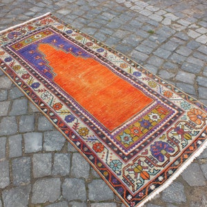 May include: A traditional Turkish prayer rug with a bright orange center and a colorful geometric border. The rug has a fringed edge and is made of wool.