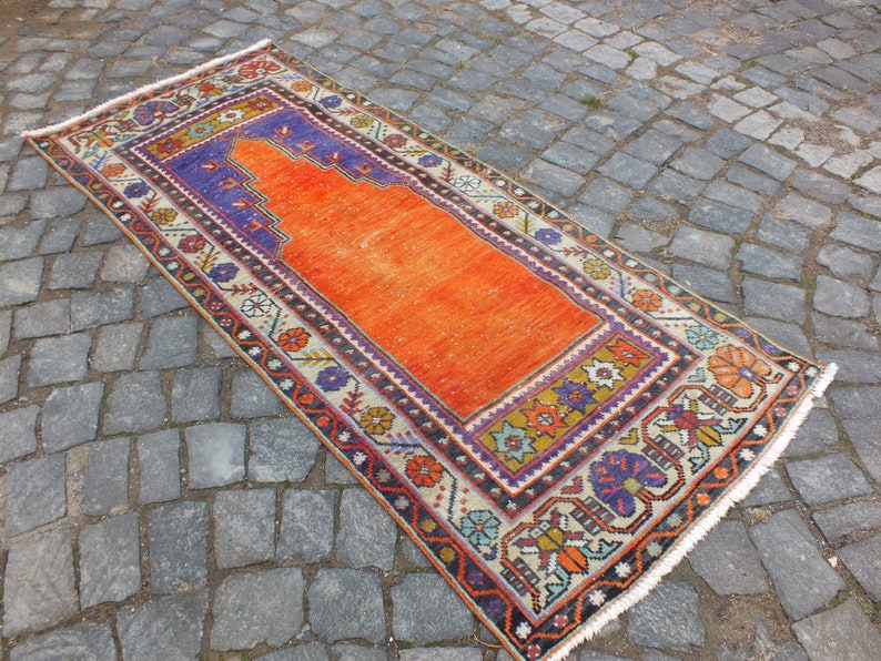 May include: A traditional Turkish prayer rug with a bright orange center and a colorful geometric border. The rug has a fringed edge and is made of wool.