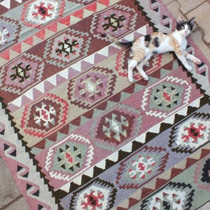May include: A rectangular, patterned rug with a geometric design in shades of pink, brown, gray, and white. The rug features repeating diamond and floral motifs. A calico cat is lying on the rug.