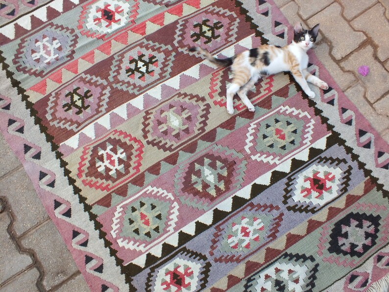 May include: A rectangular, patterned rug with a geometric design in shades of pink, brown, gray, and white. The rug features repeating diamond and floral motifs. A calico cat is lying on the rug.