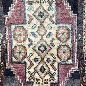 Op de afbeelding: Close-up van een rechthoekig tapijt met een centraal geometrisch ontwerp. Het tapijt heeft een cr&egrave;mekleurig midden met een ruitvormig patroon in tinten teal, roest en paars. De rand is donkerblauw en paars.