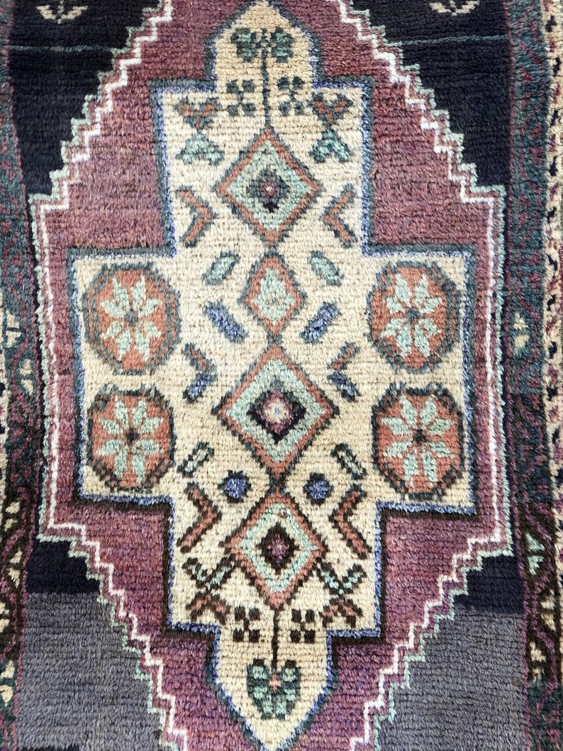 Op de afbeelding: Close-up van een rechthoekig tapijt met een centraal geometrisch ontwerp. Het tapijt heeft een cr&egrave;mekleurig midden met een ruitvormig patroon in tinten teal, roest en paars. De rand is donkerblauw en paars.