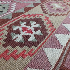 May include: Close-up of a woven rug with a geometric pattern. The rug features a combination of colors, including shades of pink, brown, beige, and white. The design includes repeating shapes and lines, creating a textured surface.