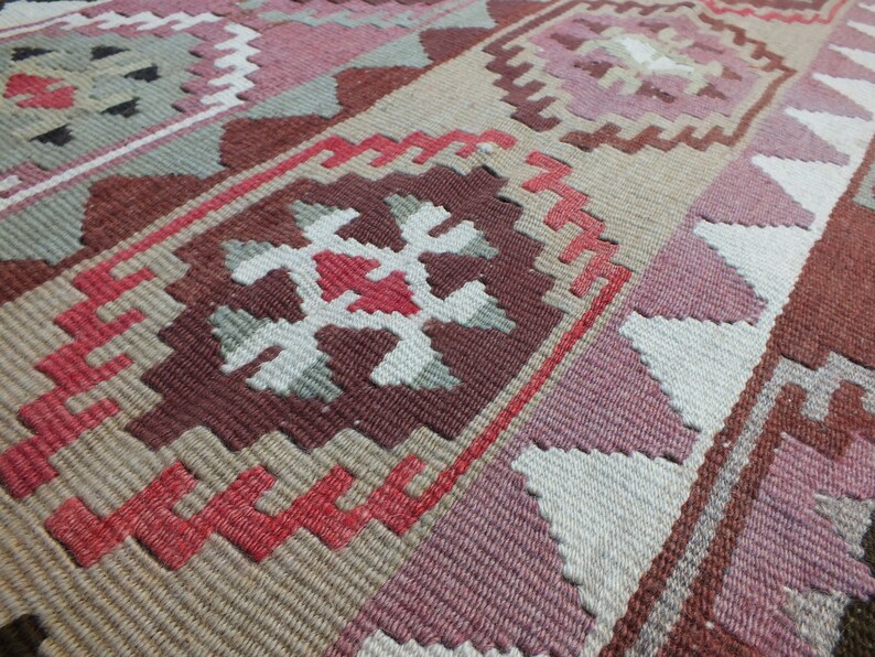 May include: Close-up of a woven rug with a geometric pattern. The rug features a combination of colors, including shades of pink, brown, beige, and white. The design includes repeating shapes and lines, creating a textured surface.
