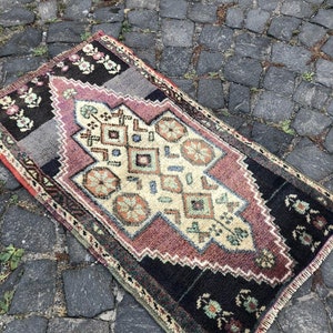 Op de afbeelding: Een rechthoekig, vintage tapijt met een centraal ruitpatroon in cr&egrave;me, mauve en teal. Het tapijt heeft een zwarte rand met bloemaccenten en een grijs gedeelte. Het tapijt ligt op een kasseienoppervlak.