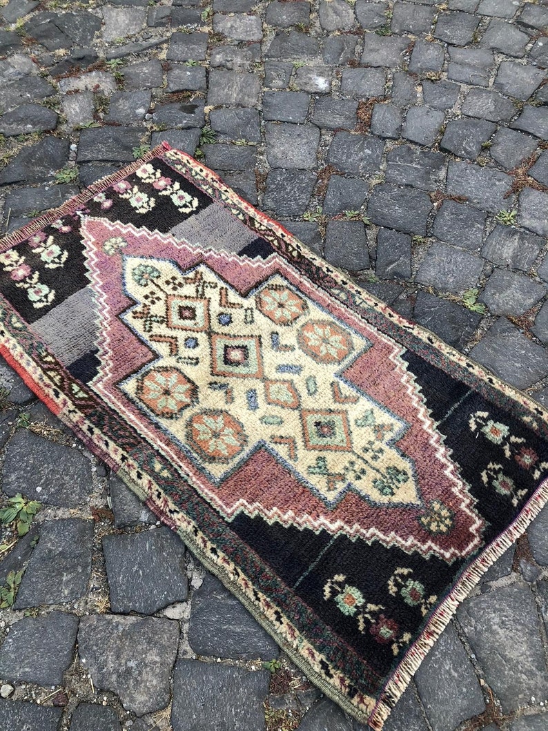 Op de afbeelding: Een rechthoekig, vintage tapijt met een centraal ruitpatroon in cr&egrave;me, mauve en teal. Het tapijt heeft een zwarte rand met bloemaccenten en een grijs gedeelte. Het tapijt ligt op een kasseienoppervlak.