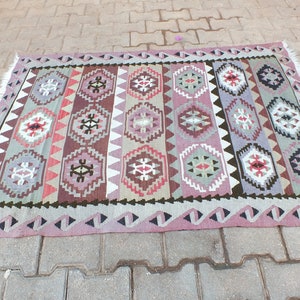 May include: A rectangular, hand-woven rug with a geometric pattern. The rug features a repeating design of diamond and hexagonal shapes in shades of pink, purple, brown, and green. The edges are finished with a fringe.