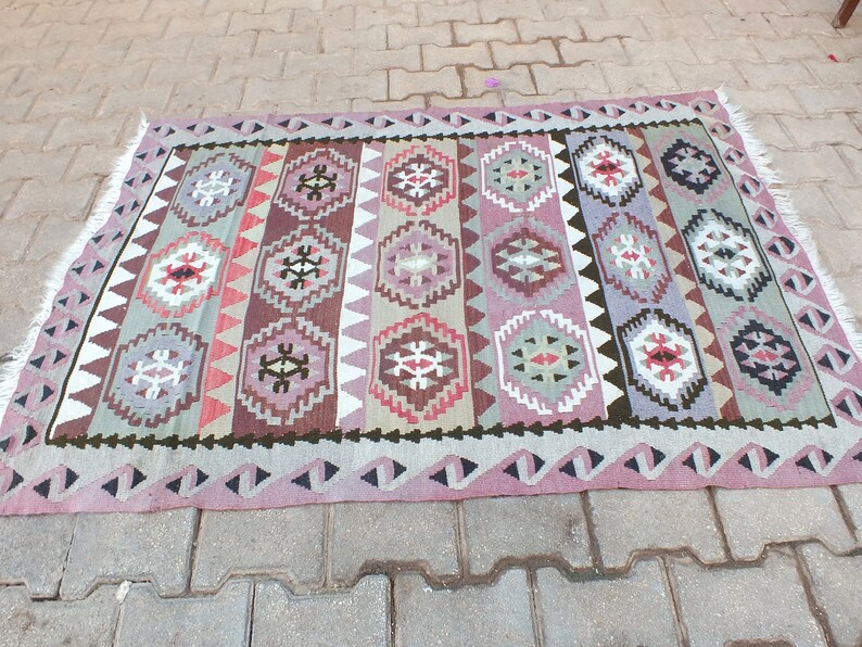 May include: A rectangular, hand-woven rug with a geometric pattern. The rug features a repeating design of diamond and hexagonal shapes in shades of pink, purple, brown, and green. The edges are finished with a fringe.