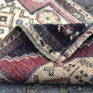 Op de afbeelding: Opgeslagen vintage tapijt met een gedetailleerd geometrisch patroon. Het tapijt heeft een mix van kleuren, waaronder bordeauxrood, cr&egrave;me, marineblauw en vleugjes teal en oranje. De randen zijn gerafeld en het tapijt lijkt van geweven materiaal te zijn.