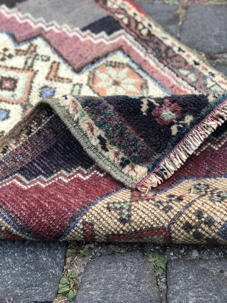 Op de afbeelding: Opgeslagen vintage tapijt met een gedetailleerd geometrisch patroon. Het tapijt heeft een mix van kleuren, waaronder bordeauxrood, cr&egrave;me, marineblauw en vleugjes teal en oranje. De randen zijn gerafeld en het tapijt lijkt van geweven materiaal te zijn.