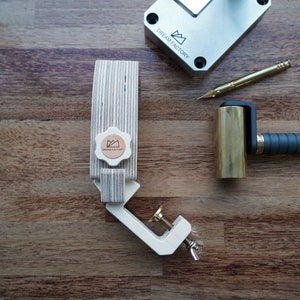 May include: A wooden clamp with a brass knob and a metal clamp. The clamp is on a wooden surface. There is a brass roller with a black handle and a metal tool with a gold tip on the surface as well. The clamp has a logo that says "Dream Factory" on it.