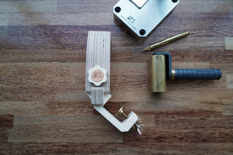 May include: A wooden clamp with a brass knob and a metal clamp. The clamp is on a wooden surface. There is a brass roller with a black handle and a metal tool with a gold tip on the surface as well. The clamp has a logo that says "Dream Factory" on it.