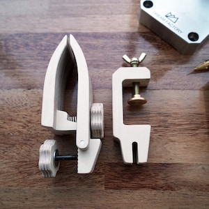 May include: Two wooden clamps for woodworking projects. The clamp on the left has a knob to tighten it, while the clamp on the right has a screw and a brass knob. The clamps are on a wooden surface.