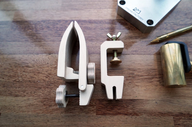 May include: Two wooden clamps for woodworking projects. The clamp on the left has a knob to tighten it, while the clamp on the right has a screw and a brass knob. The clamps are on a wooden surface.
