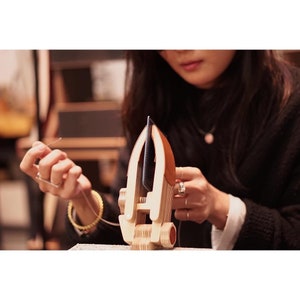 May include: A wooden craft project with a black leather piece attached. The project is shaped like a small boat with a rounded bottom and a pointed top. The person is holding a needle and thread in their left hand and is working on the project with their right hand.