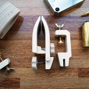 May include: Three wooden clamps with brass accents, a brass hammer with a black handle, and a metal block with the text "FACTORY" on it. The clamps are on a wooden surface.